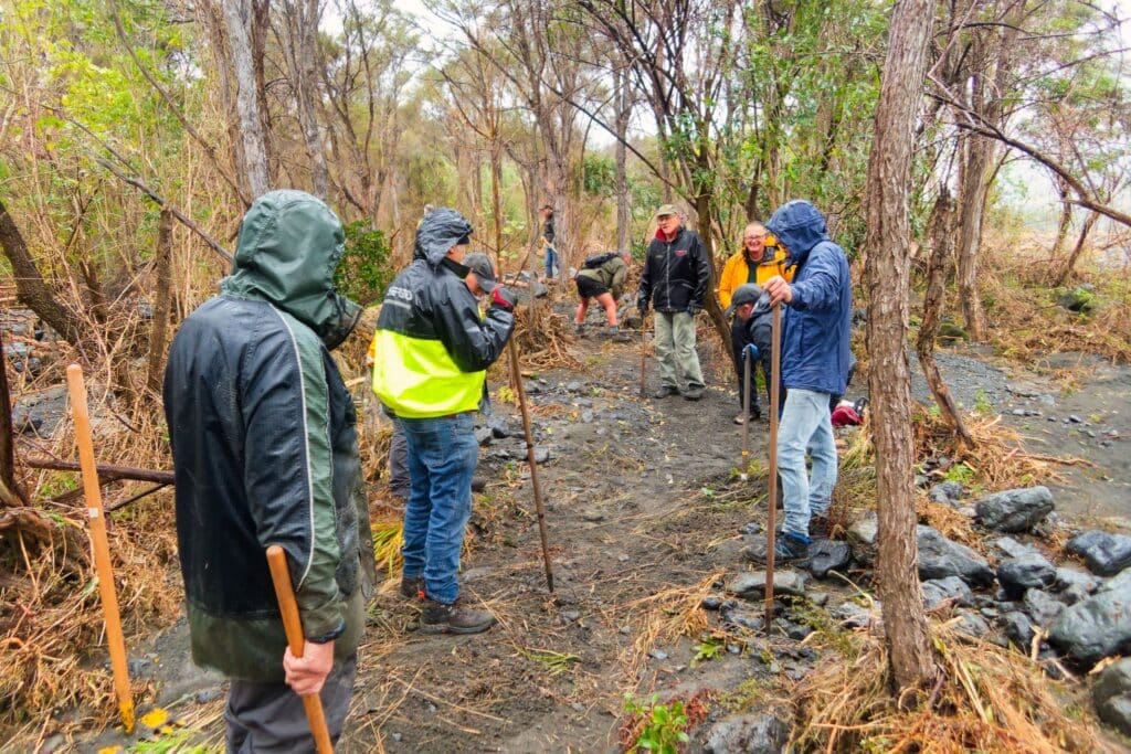 Group of hikers in rain gear with walking sticks on a muddy, rocky forest path.
