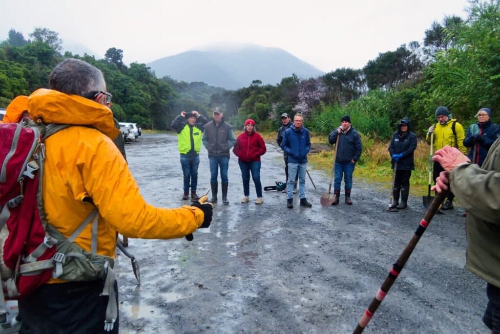 Group of hikers in a wet gravel area with mountains and trees in the background.