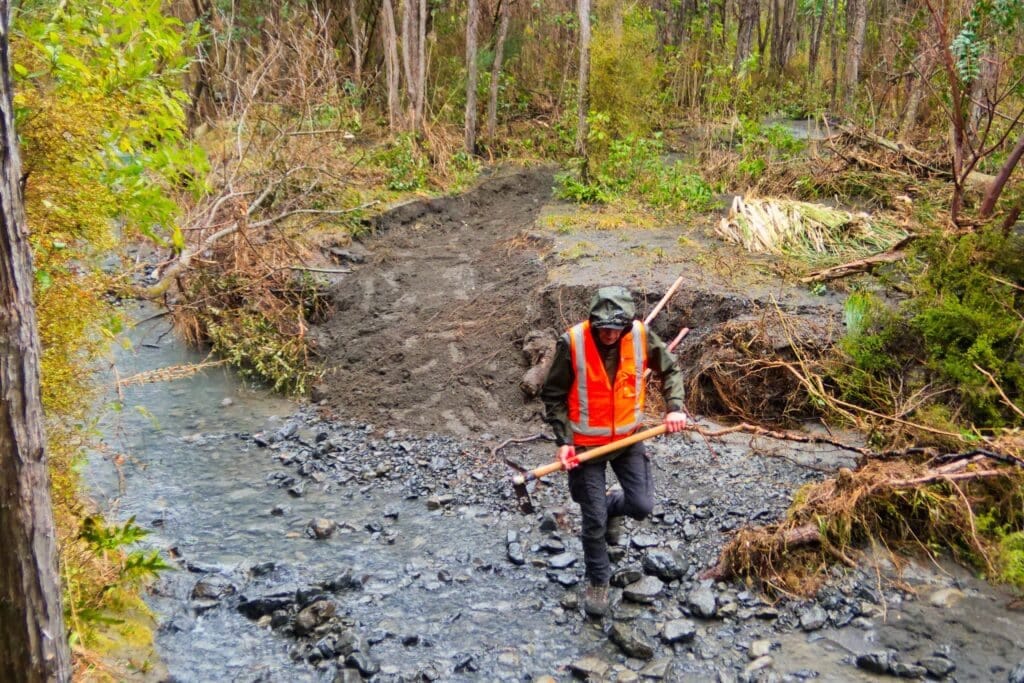 Person in orange safety vest walking through a muddy rocky riverbed in a forest.