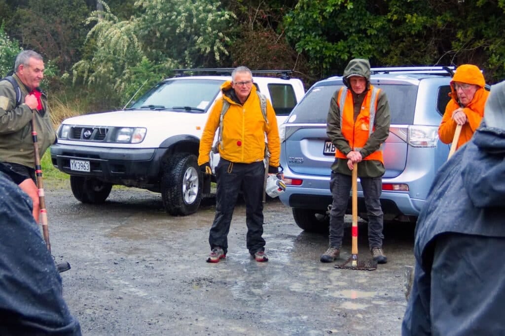 Group of men in outdoor gear with shovels beside parked SUVs on a muddy area.