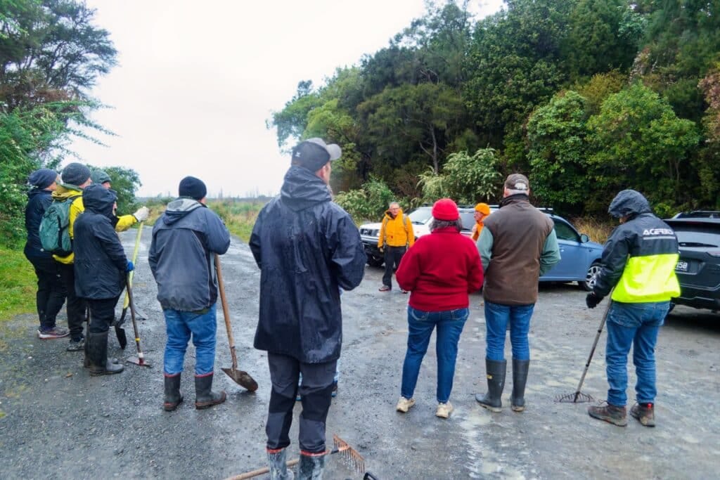 Group of people in outdoor gear standing on a damp gravel road, some with tools, near parked cars.