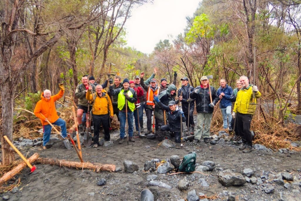 Group of people in outdoor gear with tools standing on rocky riverbank, trees in background.