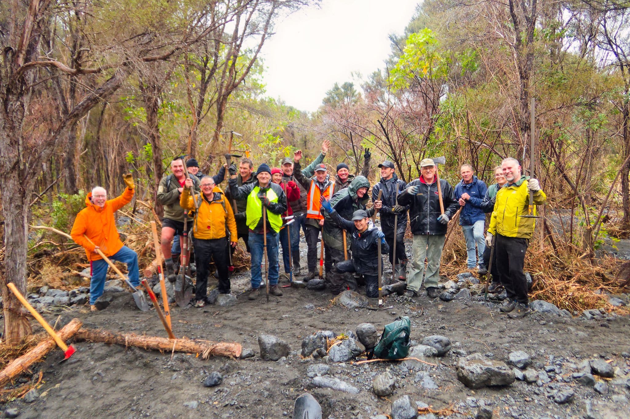 Group of people in outdoor gear with tools standing on rocky riverbank, trees in background.