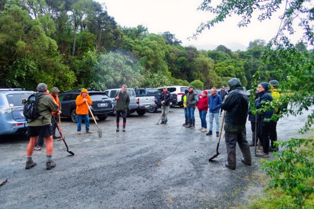 Group of people in outdoor gear with shovels in a gravel parking area, surrounded by trees.