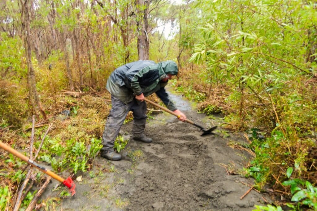 Person in a green rain jacket shoveling mud on a forest trail.